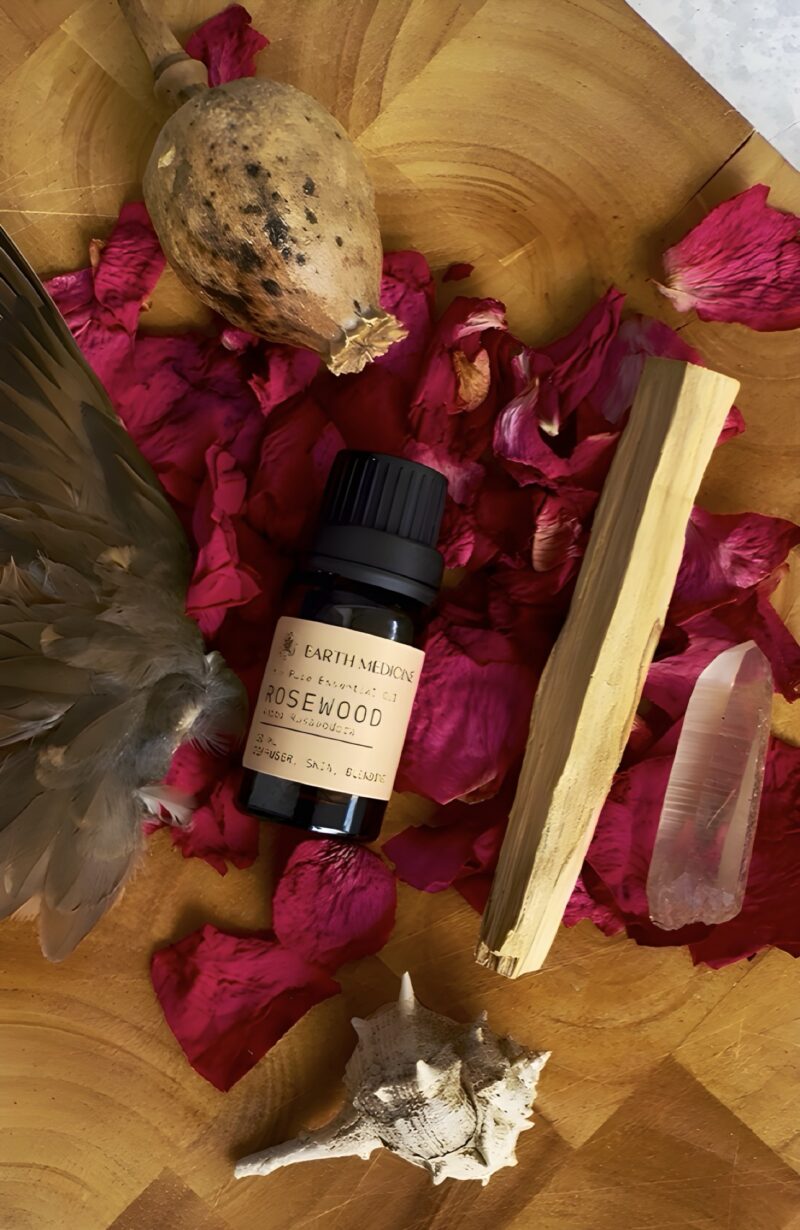 Rosewood essentiële olie op een houten plateau, omringd door roze bloemblaadjes, palo santo, en veren – een zachte en spirituele compositie in aardse tinten.