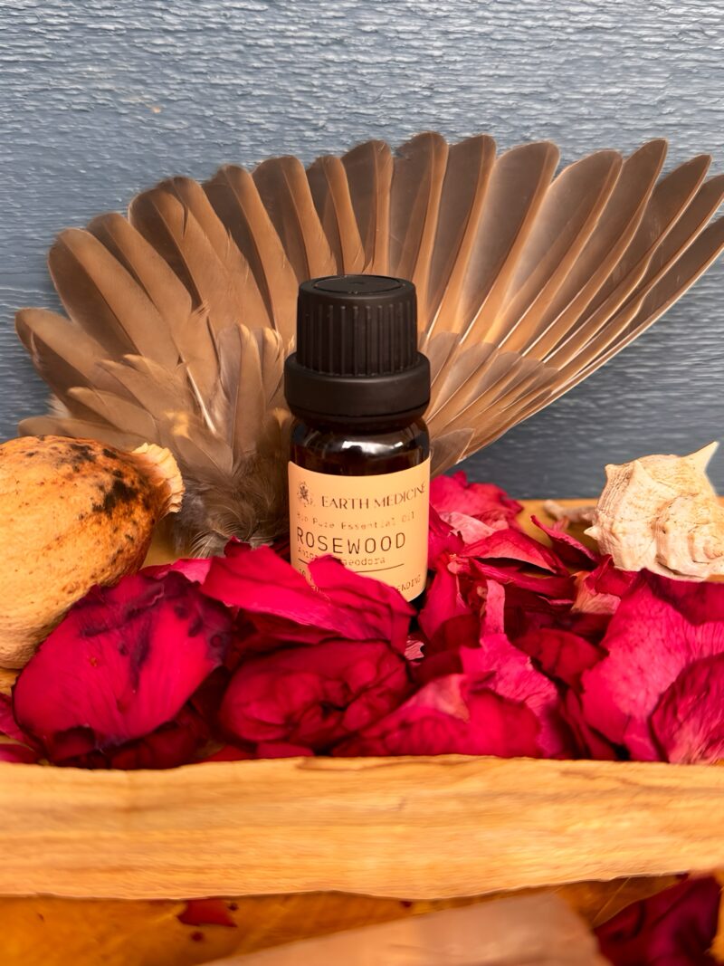 Rosewood essentiële olie op een houten plateau, omringd door roze bloemblaadjes, palo santo, en veren – een zachte en spirituele compositie in aardse tinten.